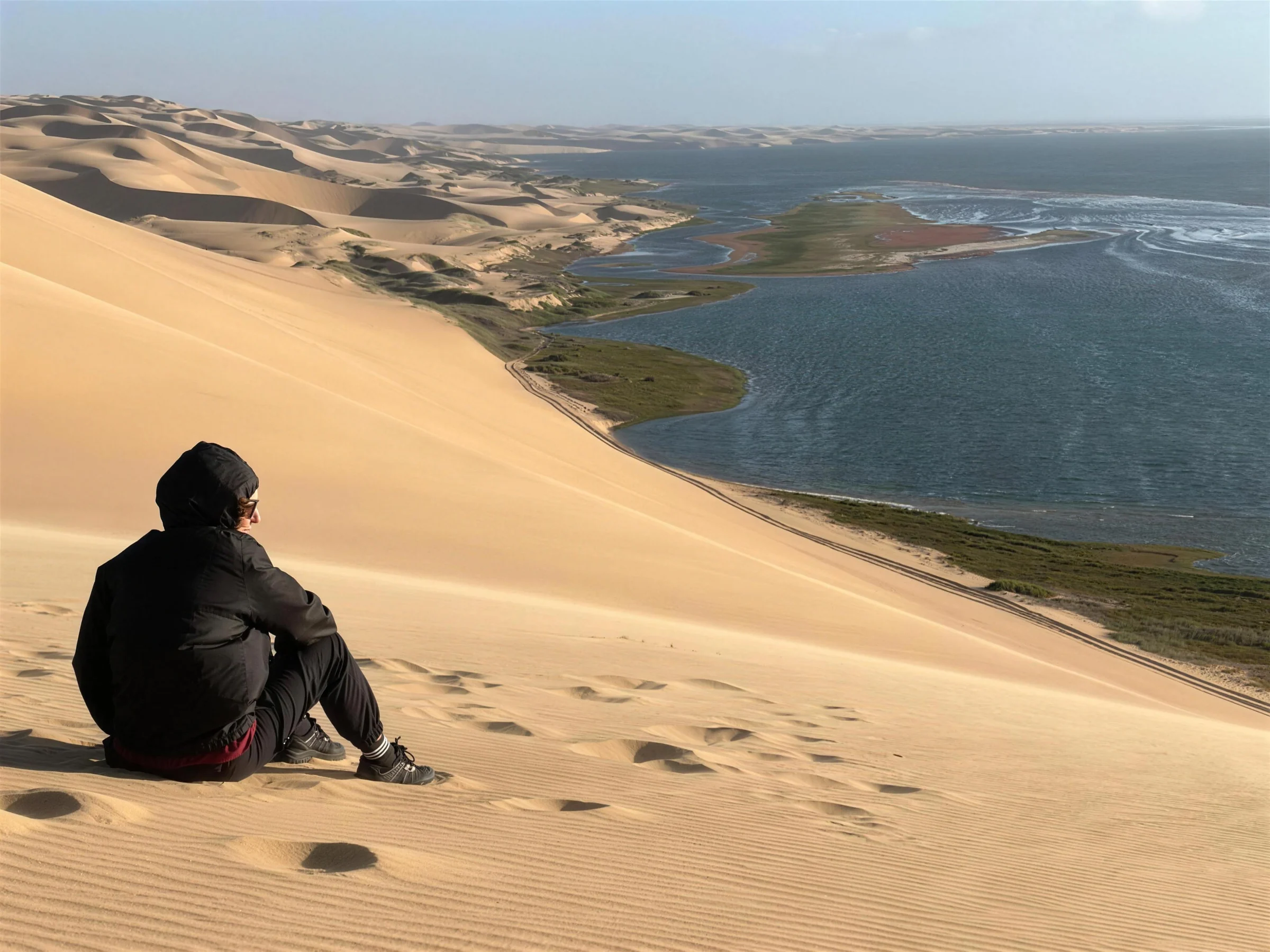 A person seated high on a Sandwich Harbour dune above coastal water